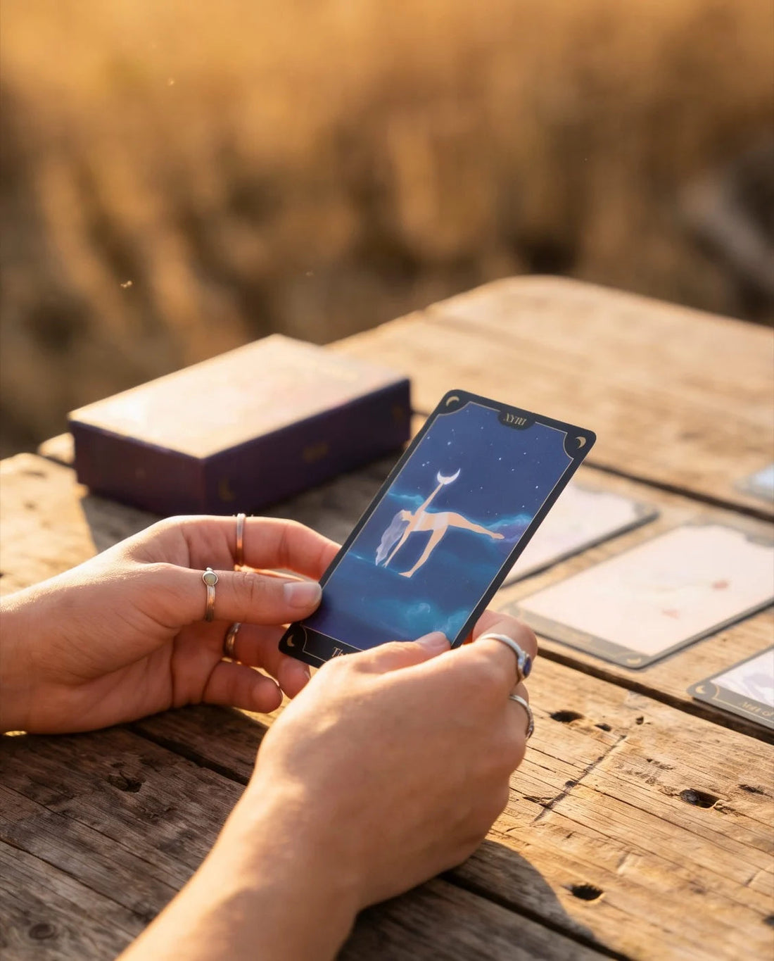 Hands holding The Moon card at a wooden table in golden hour light